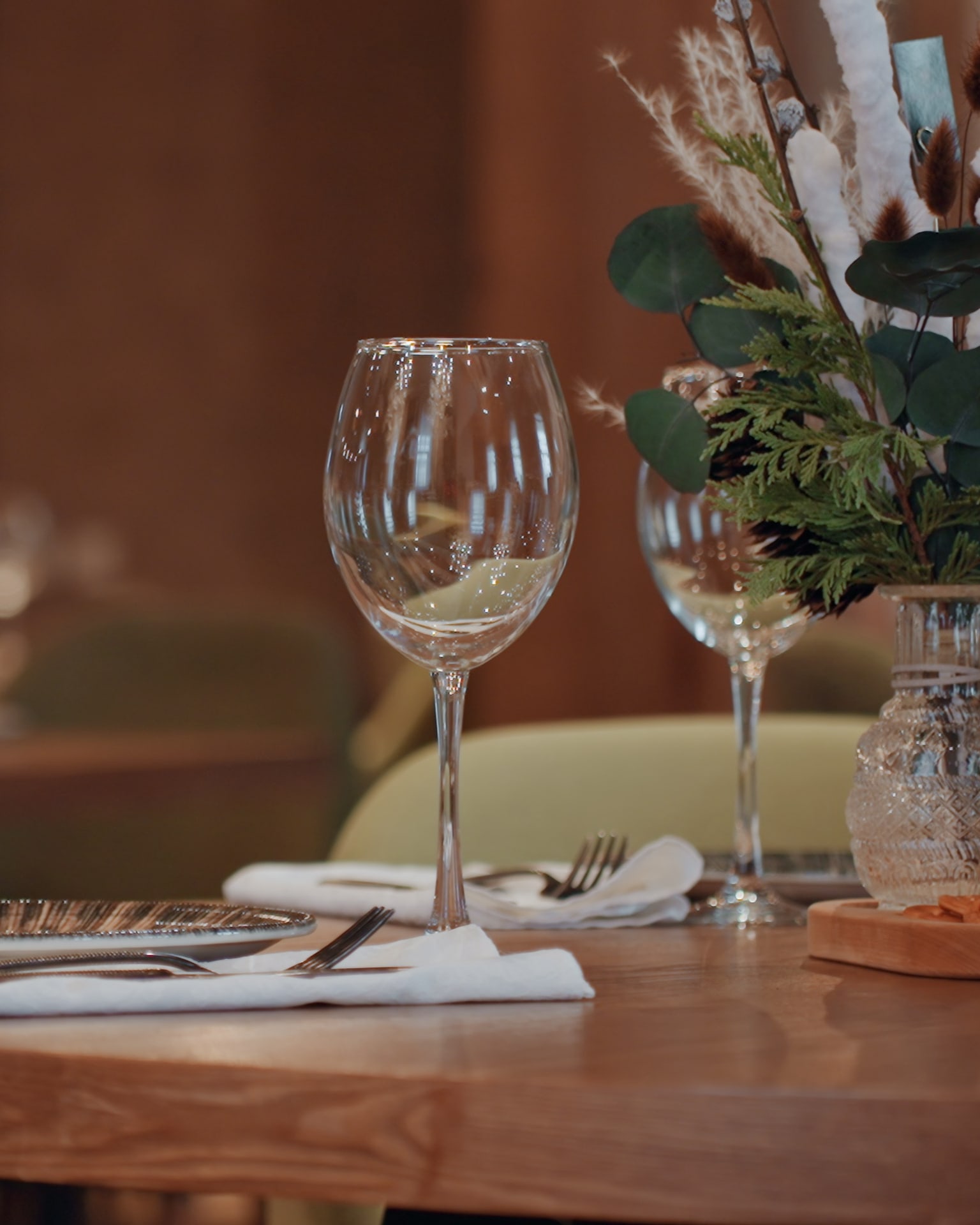 A small timber dining table set with linen, glassware and a vase of foliage