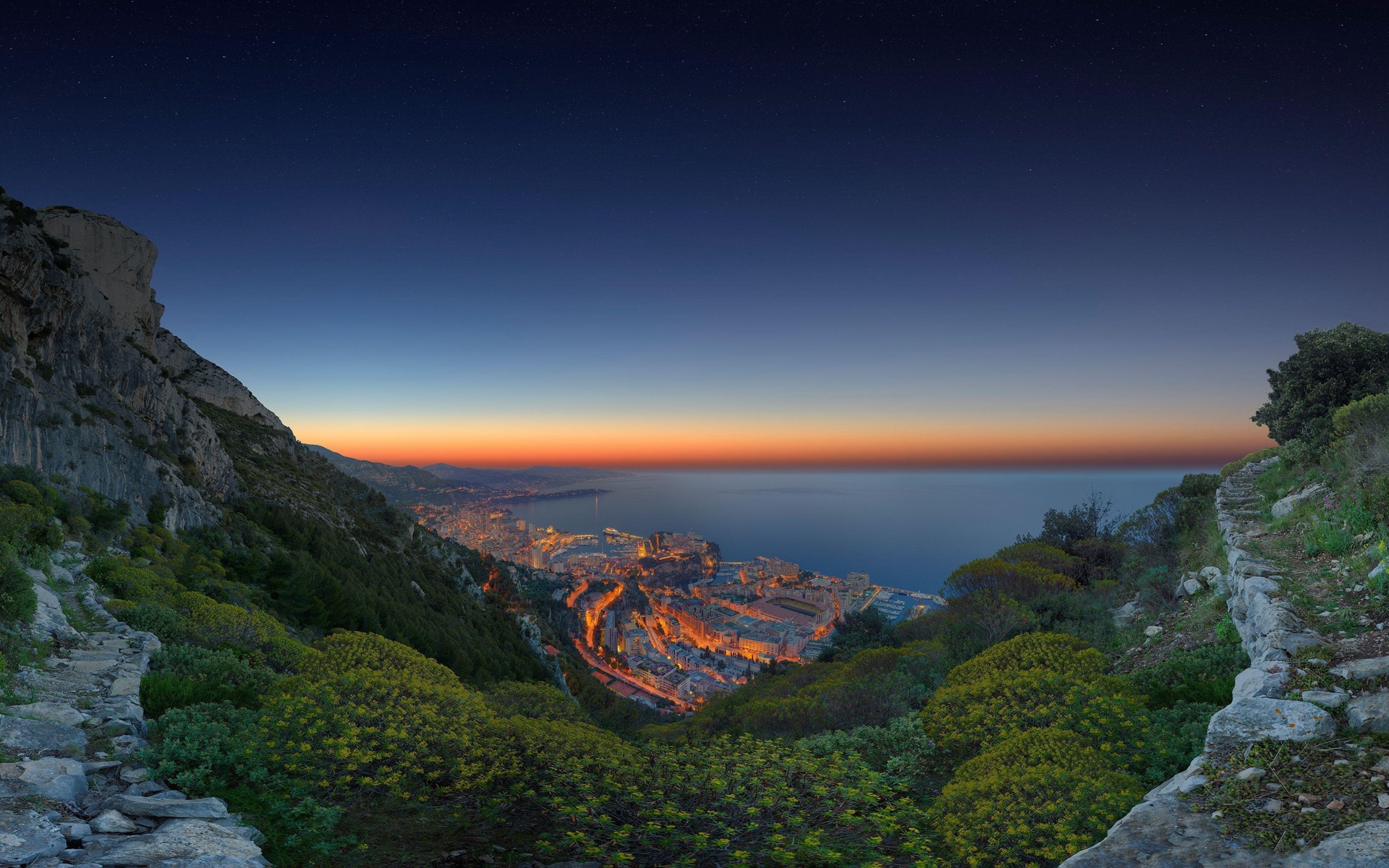 A valley path descending toward a lit coastal city at dusk